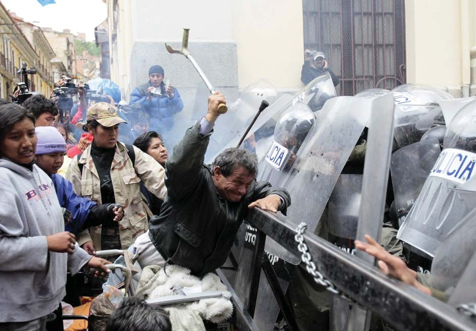 A man with several people behind him uses a walking stick to hit a wall of riot cops holding shields.