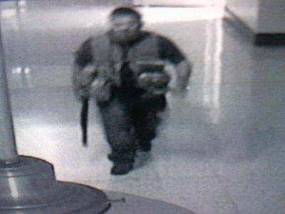 Still from a ecurity camera showing a man in a utility vest walking around with a shotung at his side.