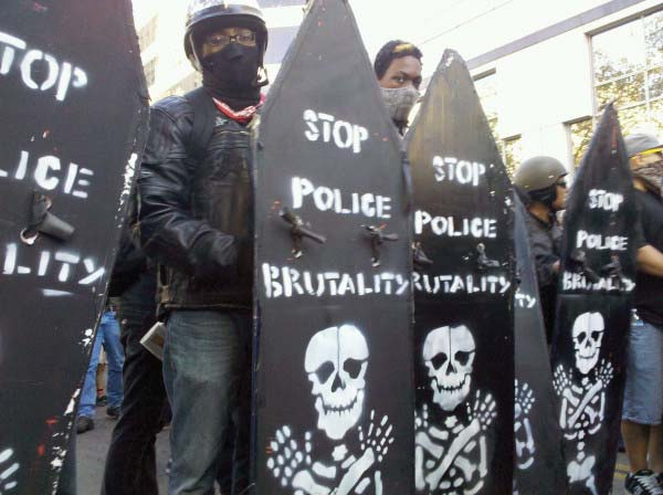 Black protesters with masks stand behind a wall of homemade shields with 